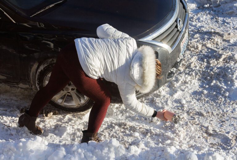 A driver uses a coffee mug to dig snow out from in front of the wheels of their vehicle.