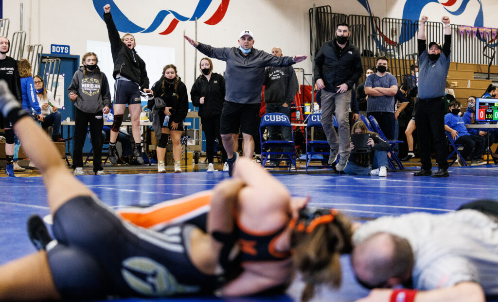 Meridian coaches and wrestlers cheer from the sidelines as Mirage Deneal struggles with an opponent on the wrestling mat.