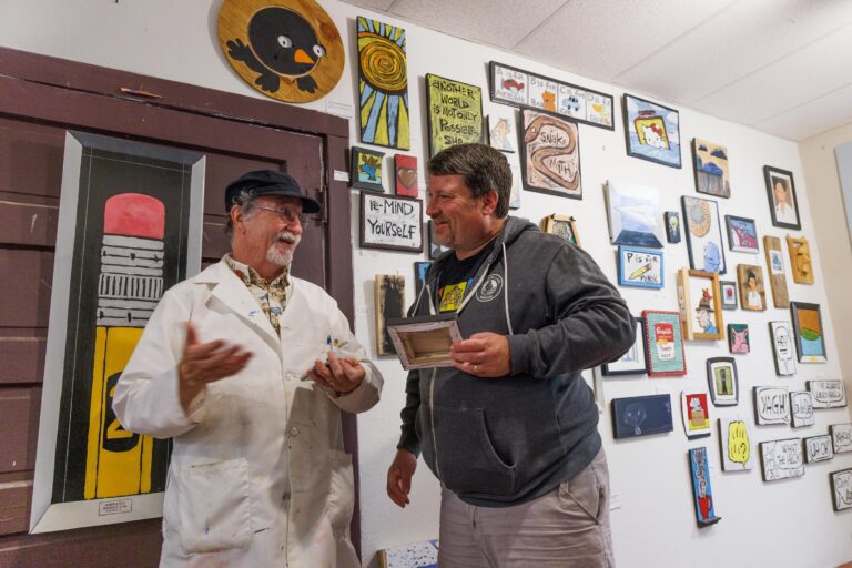 Artist Frank Frazee talks with Erik Eliason while surrounded by multiple framed art pieces.