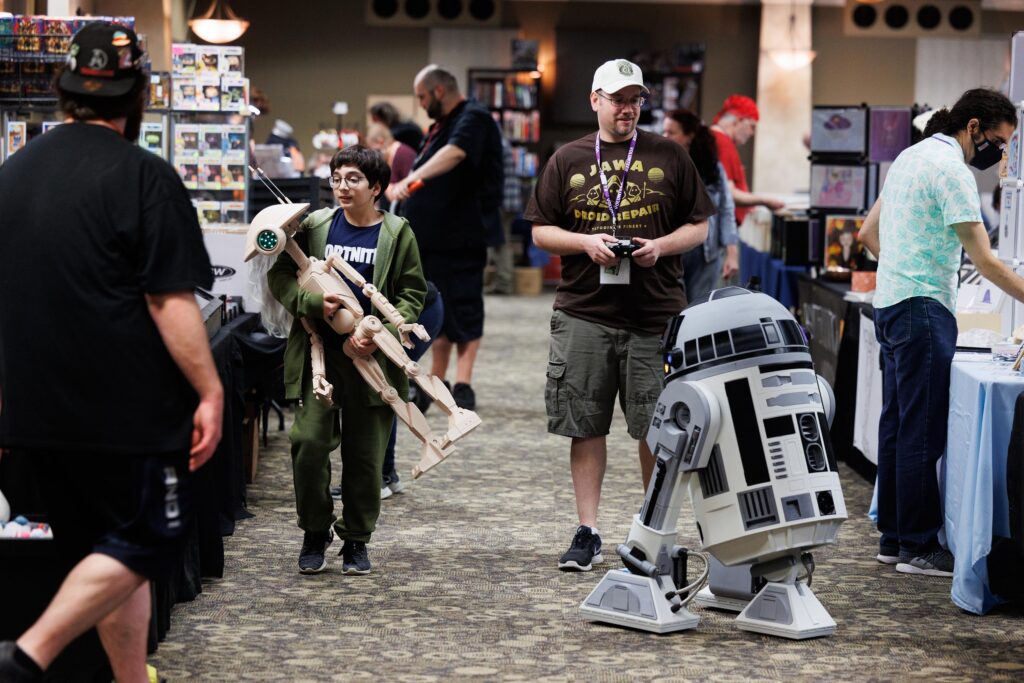 David Wolf operates an R2-D2 robot while his son, Noah Wolf, carries a worker robot named Bob around the Bellingham Comicon as other attendees browse artist alley booths.