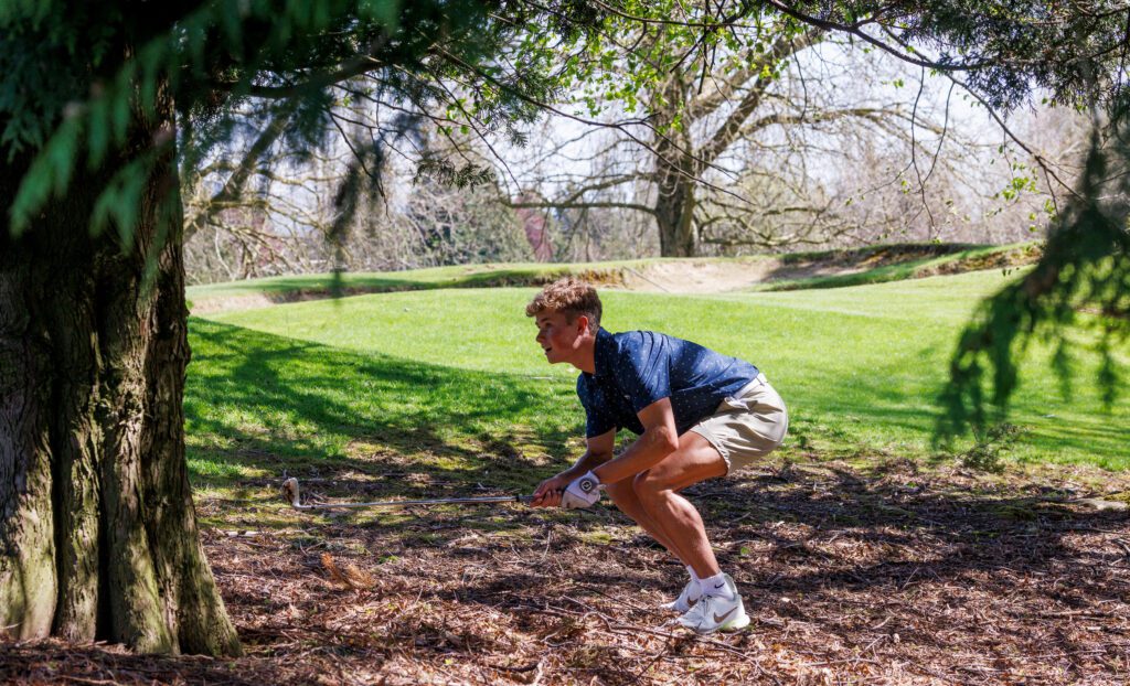 Lynden Christian's Griffin Dykstra kneels with his golf club to watch where the golf ball is heading.