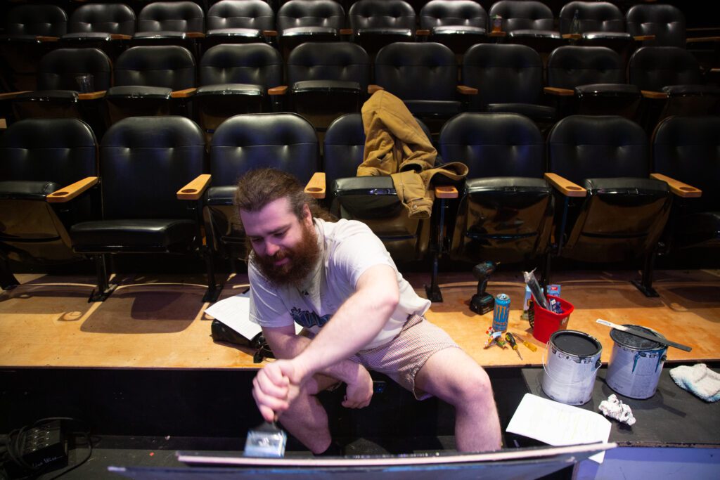 Tyler Detrick paints a set piece at the Sylvia Center in front of the audience seats.