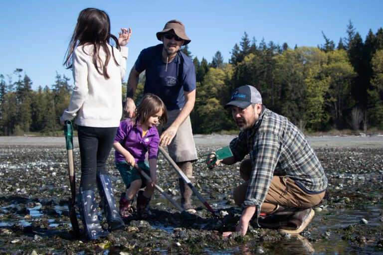 On the right, Ficus Chan of Crab Bellingham gives clamming tips while squatting close to the ground.