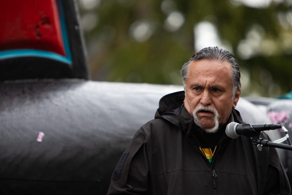 Jewell James, a Lummi carver, speaks to the crowd as light rain pours down.