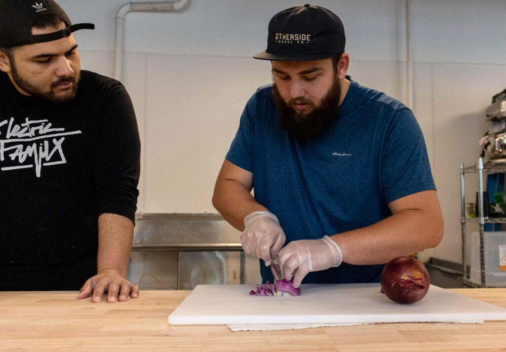 Otherside Bagel Co. chef Zach Leinan shows employee Dane Johnson the best way to slice onions.