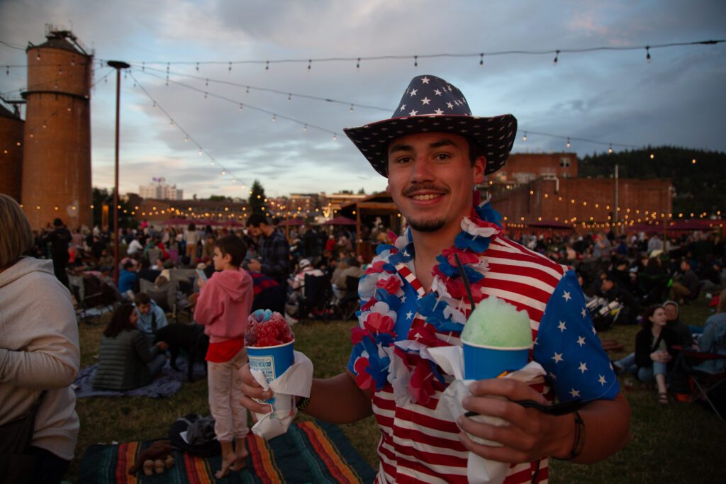 Decked out in Fourth of July gear, Hunter Ivie holds two snow cones of different flavors as he is surrounded by crowds of people.