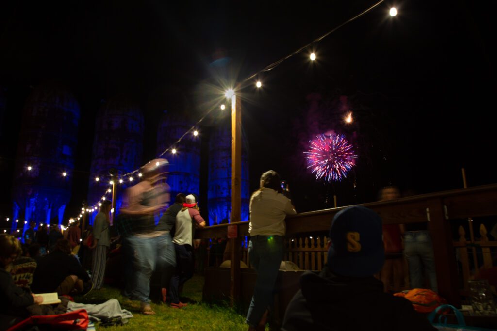 People watch fireworks light up the sky from the beer garden as some sit on the grass to watch while others lean on the wooden counters to take pictures.