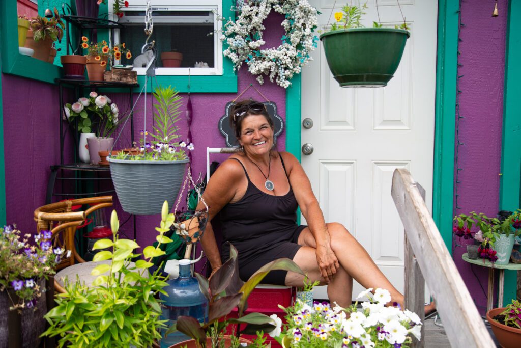 Resident and village manager Tina Hayes sits on the front porch of her purple tiny home with a big smile amongst the planters and flowers.