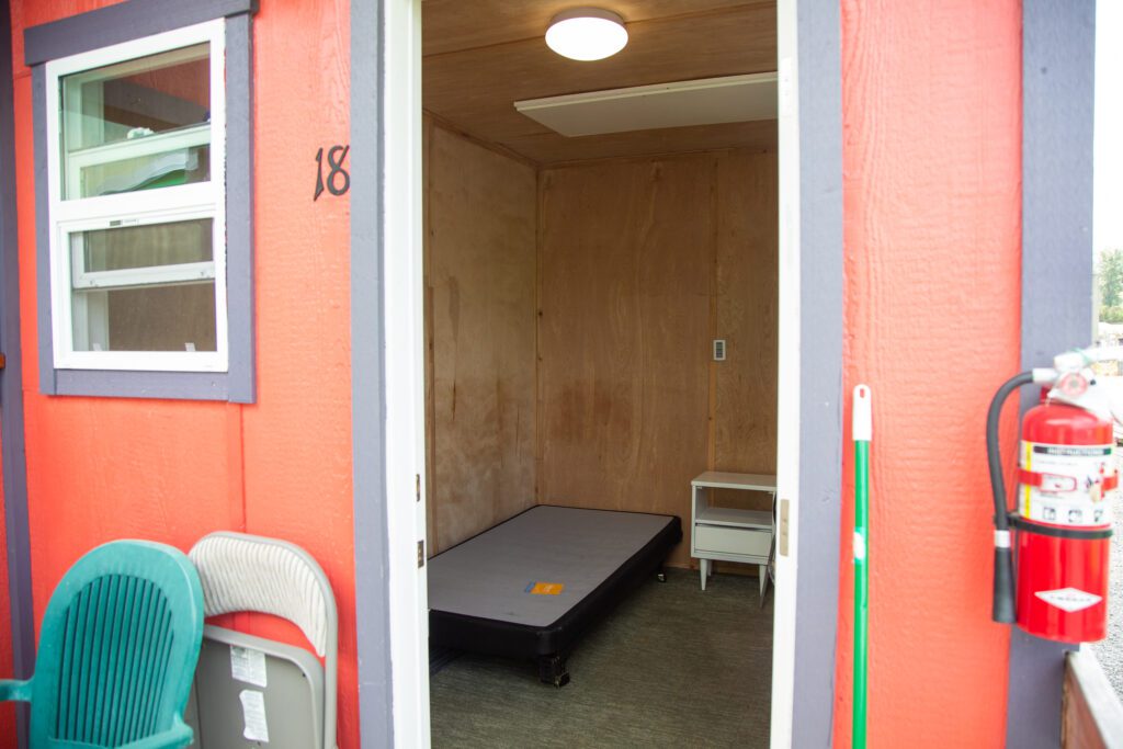 An available tiny home comes with a bed, bedside table, electrical outlets, and chairs in front of the entrance.