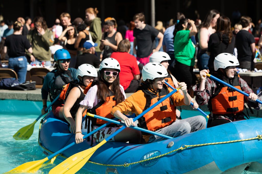 Students ride a whitewater raft a with safety gear and rows as other students chat amongst the crowd.