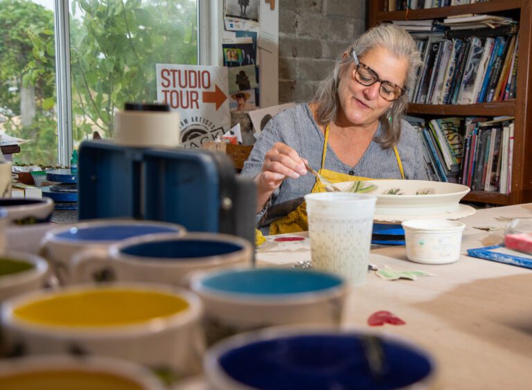 Ann Marie DeCollibus works on a plate in her home studio with multiple various coloring paints placed on the table.