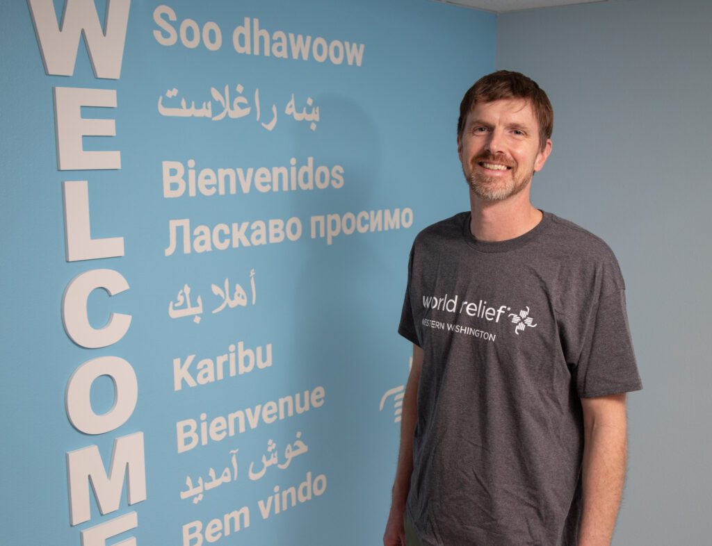 Steven Shetterly stands next to a blue wall with the word "welcome" written in different languages.