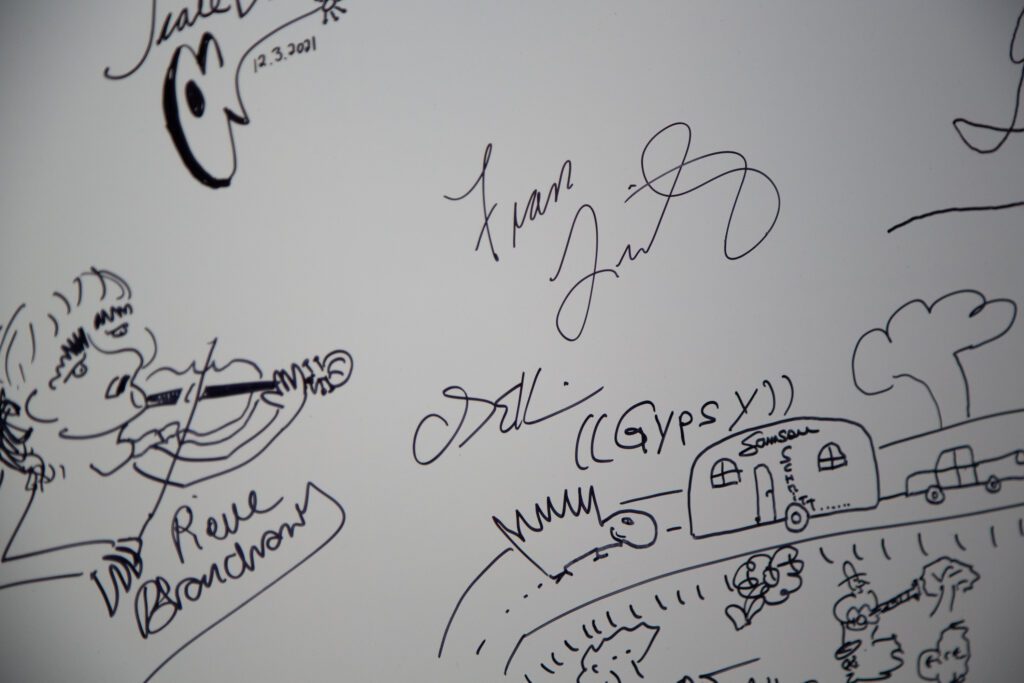 Past performers sign on a whiteboard backstage of the theater with doodles around the signatures.