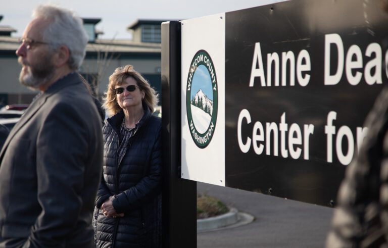 A woman in a black puffy jacket and sunglasses stands next to a sign that reads "Whatcom County Washington."