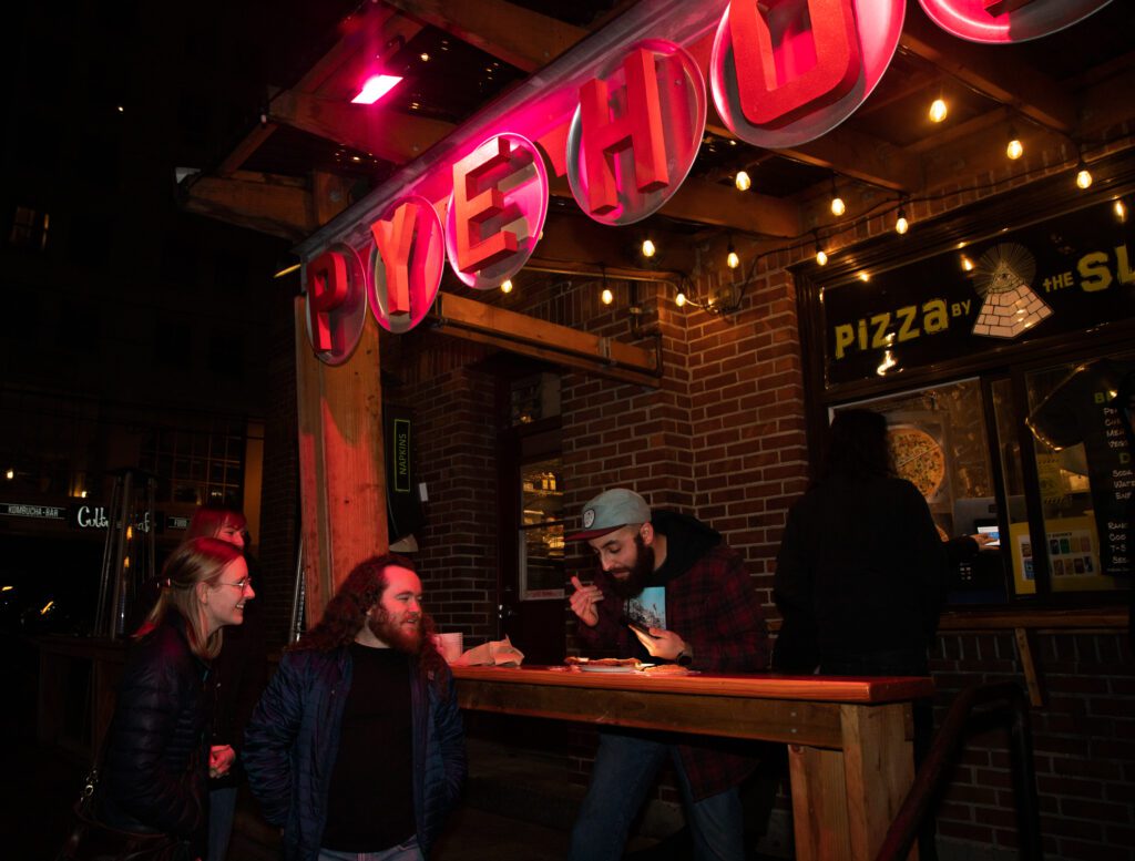 Food writer Mark Saleeb and friends inspect a fresh slice of veggie pizza underneath the bright neon lights.