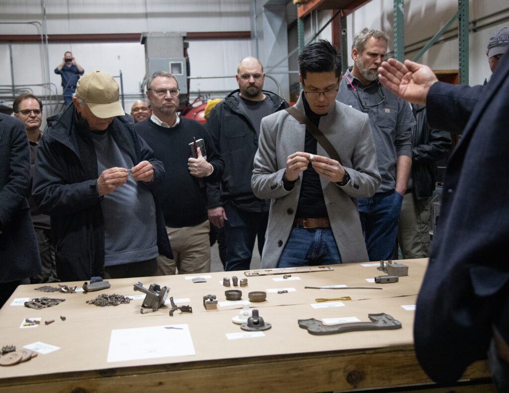 Guests on a tour of 3DX Industries view 3D-printed metal parts closely as they are laid out on a table for attendees to inspect.