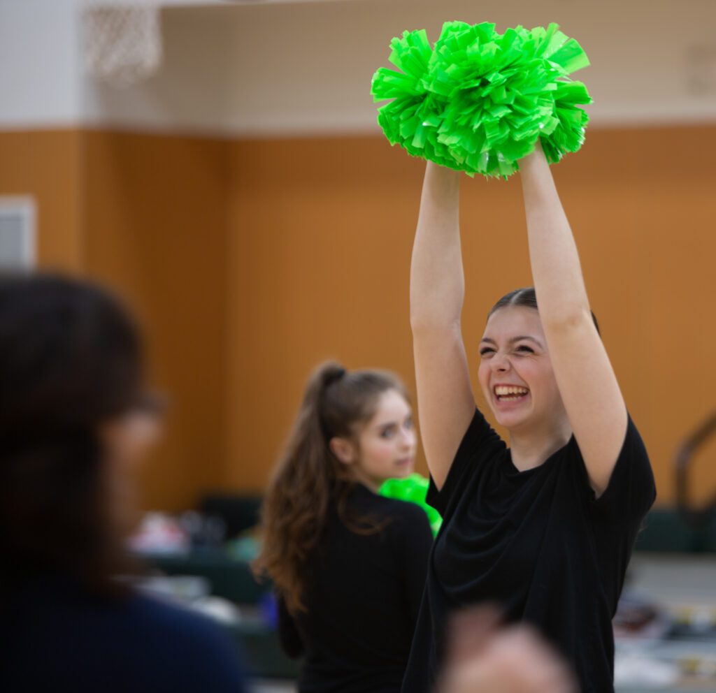 Jessie McDonald laughs with both her arms in the air holding pompoms while receiving instructions from coach Selena Arciniega-Storey.