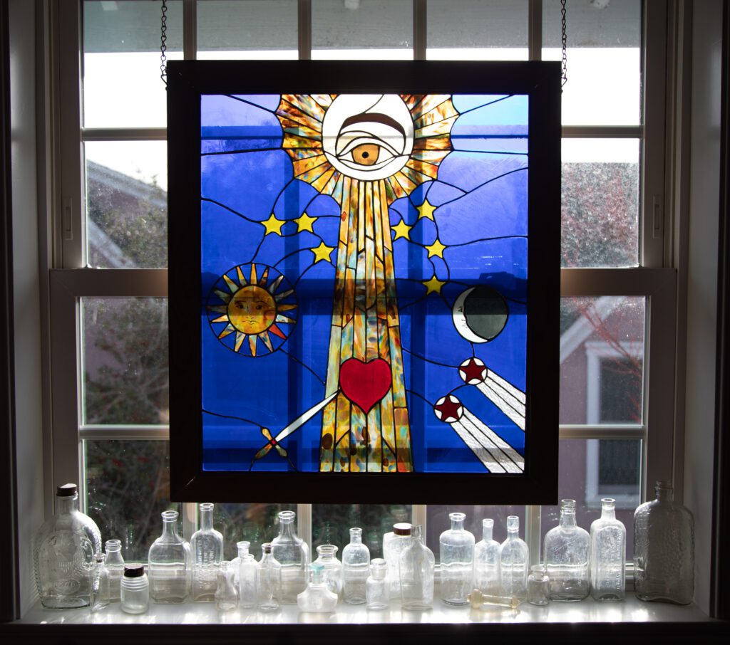 A stained glass piece over glass bottles sitting on the window sill.