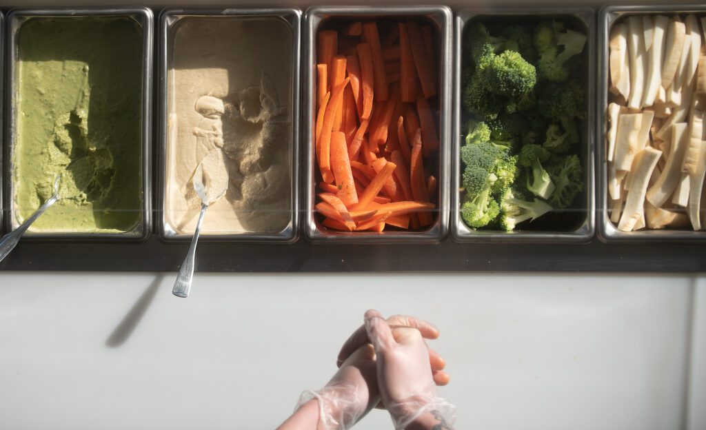 A pair of hands adjusts the plastic gloves on them near trays full of vegetables and hummus choices.