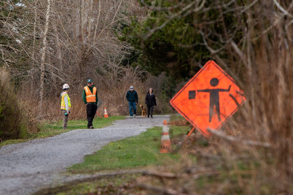 A bright orange sign warns visitors of construction work as some visitors are walking towards flaggers waiting for their next load of dirt.