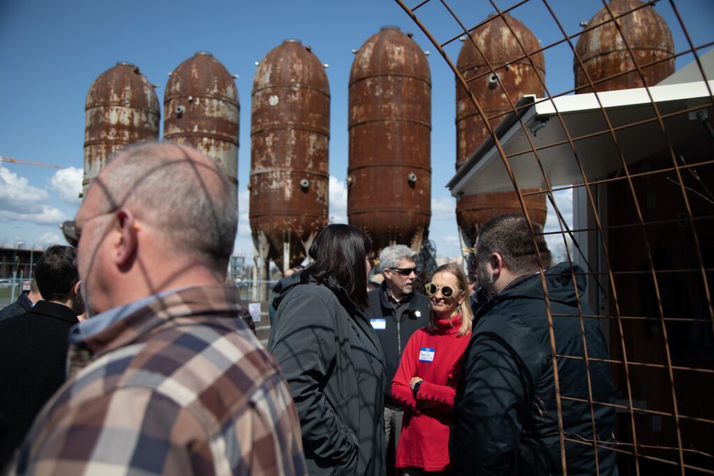 People chat and enjoy food at Rain or Shine Riviera Club with the rusty metal silos looming behind them.