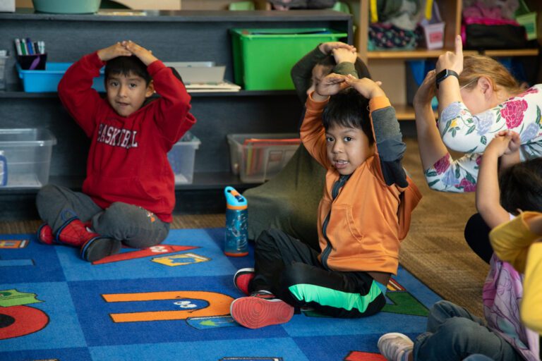 Transitional Kindergarten students participate by holding their hands over their head on the carpeted floor.