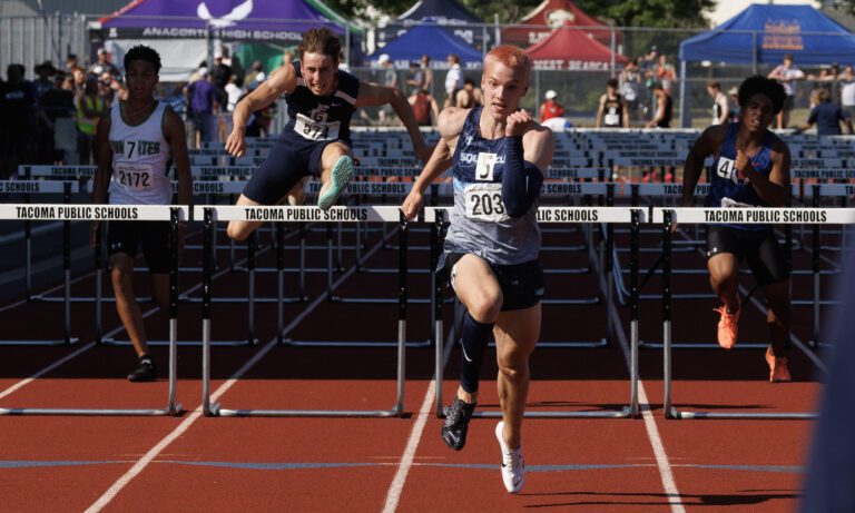 Andre Korbmacher races to the finish line as another runner jumps a hurdle behind him.