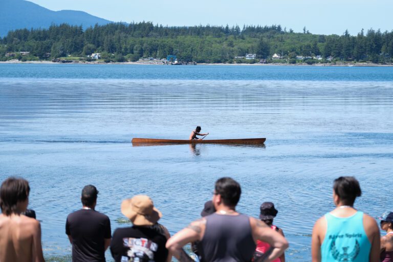 A solo kayaker pushes to the finish line as attendees watch from the shore.