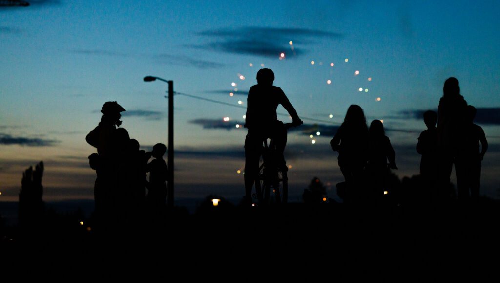 Fireworks go off in the sky as children of varying ages watch from afar, some on their bikes.