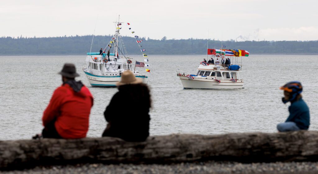 Visitors sit on large driftwood on the beach as boats idle side by side.