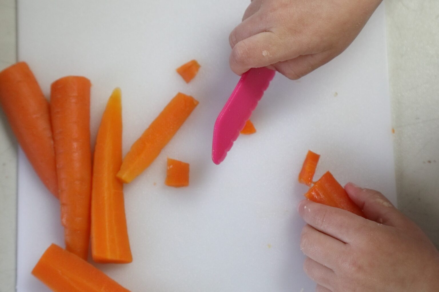 A top-down view of a child cutting carrots with a pink child-safe knife.