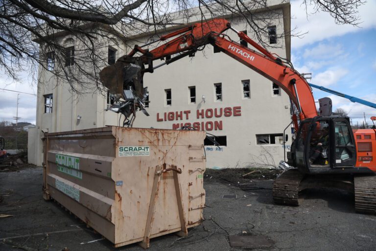 An excavator drops scrap metal into a bin in front of the lighthouse.