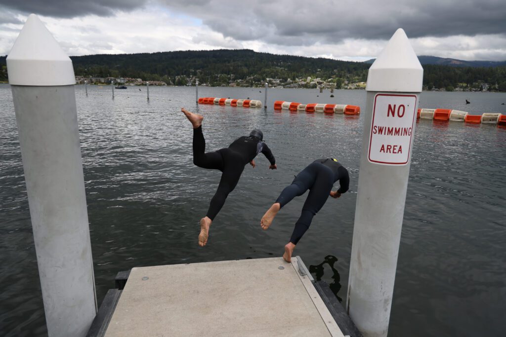 Ethan Hunger and Jake Birnel dive into Lake Whatcom in full diving suits as clouds cover the sky above.