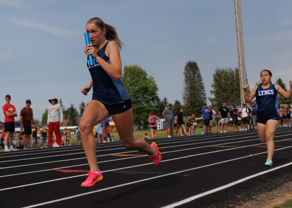 Lynden Christian's Sydney Terpstra takes the baton from teammate Morgan Delgato as spectators watch from the sidelines.