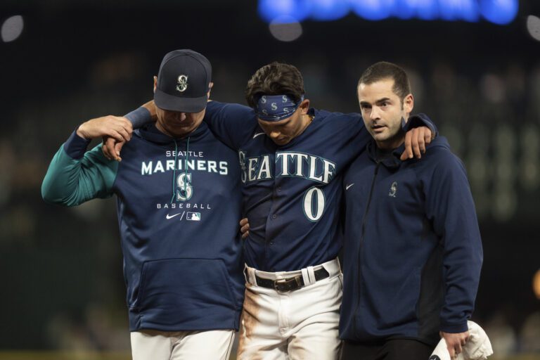 Seattle Mariners’ Sam Haggerty, center, is helped off the field with his arms around the helpers.