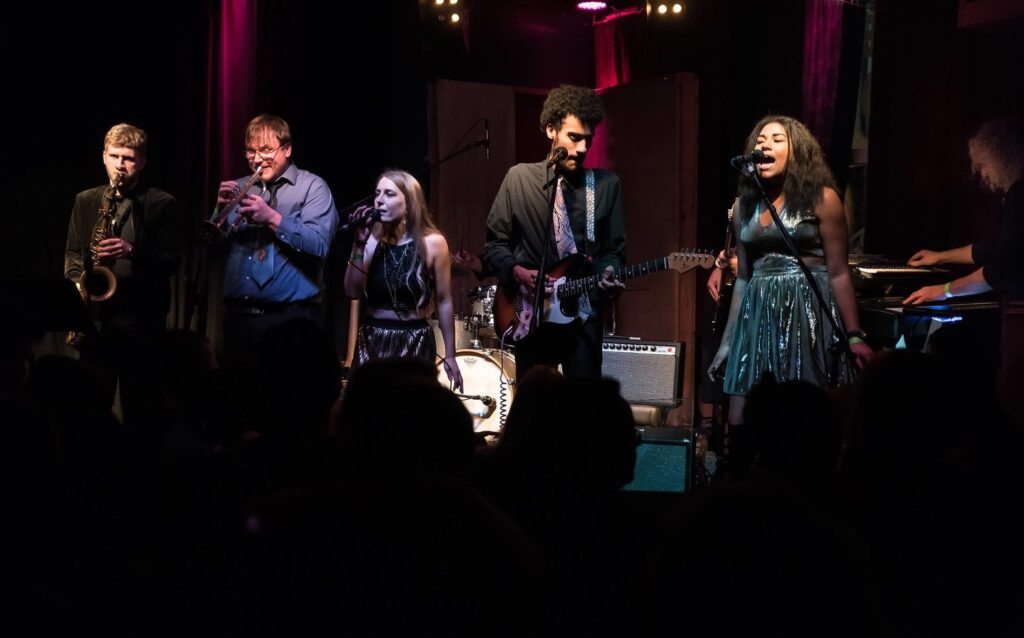 Band Baby Cakes performing on stage with their respective instruments.