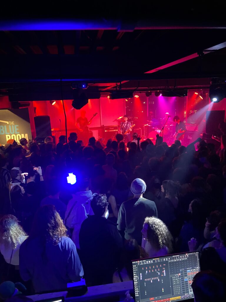 The Blue Room filled with a crowd as a band performs on stage underneath a red light.