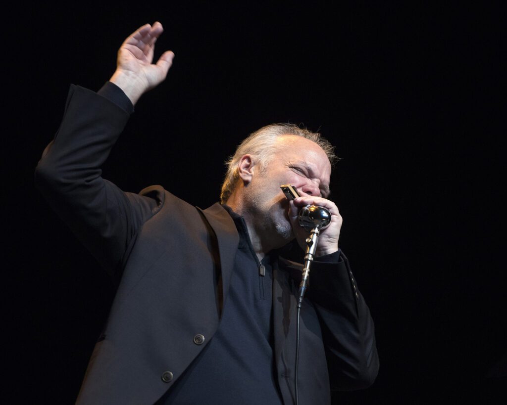 Award-winning blues vocalist, instrumentalist and songwriter Curtis Salgado plays the harmonica into the mic as he raises one arm.