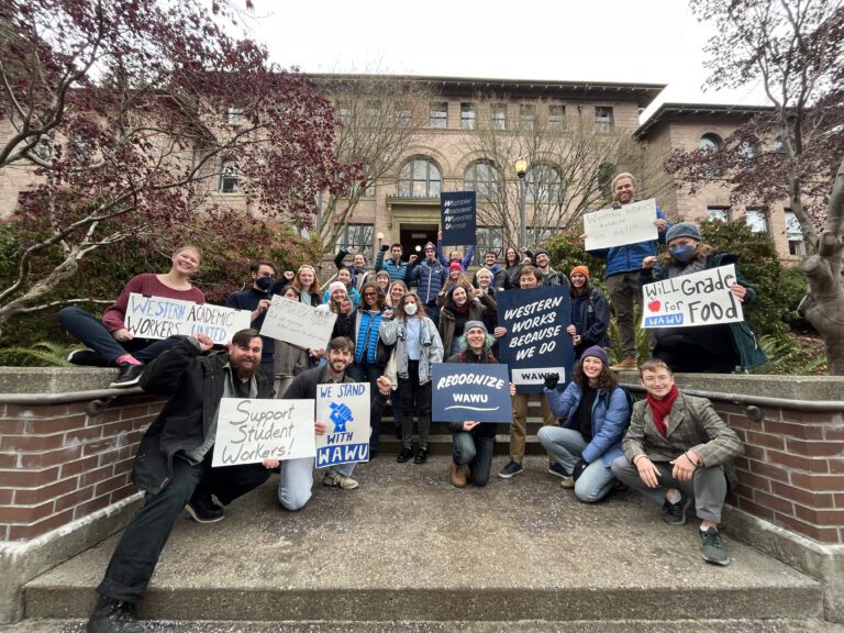 Academic Student Employees stand on the stairs of Old Main at Western Washington University with signs calling for action.