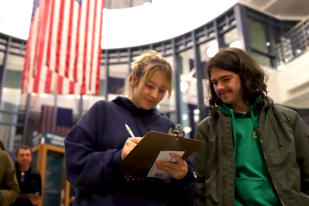 Alyssa Coleman writing on a clipboard and Tucker Zucati watches from the side as the american flag hangs behind them.