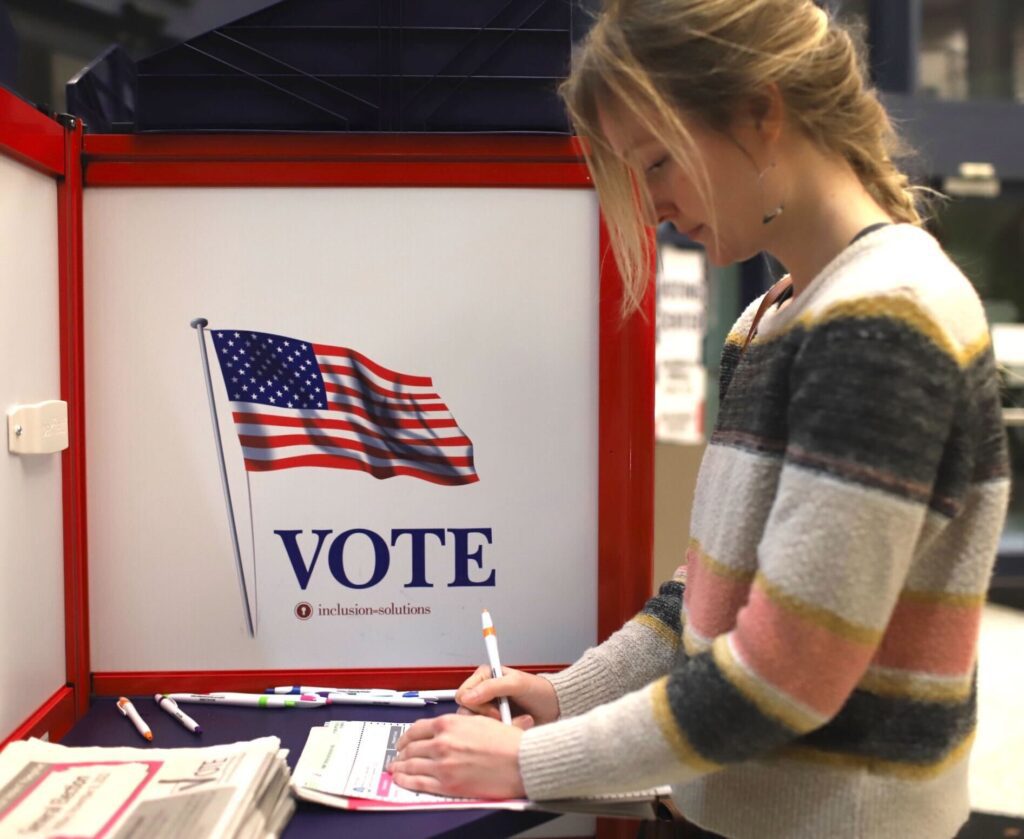 Morgan Harrington fills out her ballot in a privacy box next to an American flag above the word "VOTE".