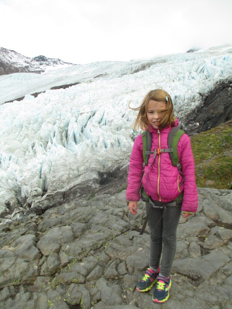 A girl takes a photo with the Coleman Glacier as wind sweeps her hair to the side.