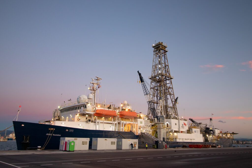 The Joint Oceanographic Institutions for Deep Earth Sampling (JOIDES) Resolution sits idle at a dock with the sun set streaking the sky behind it.