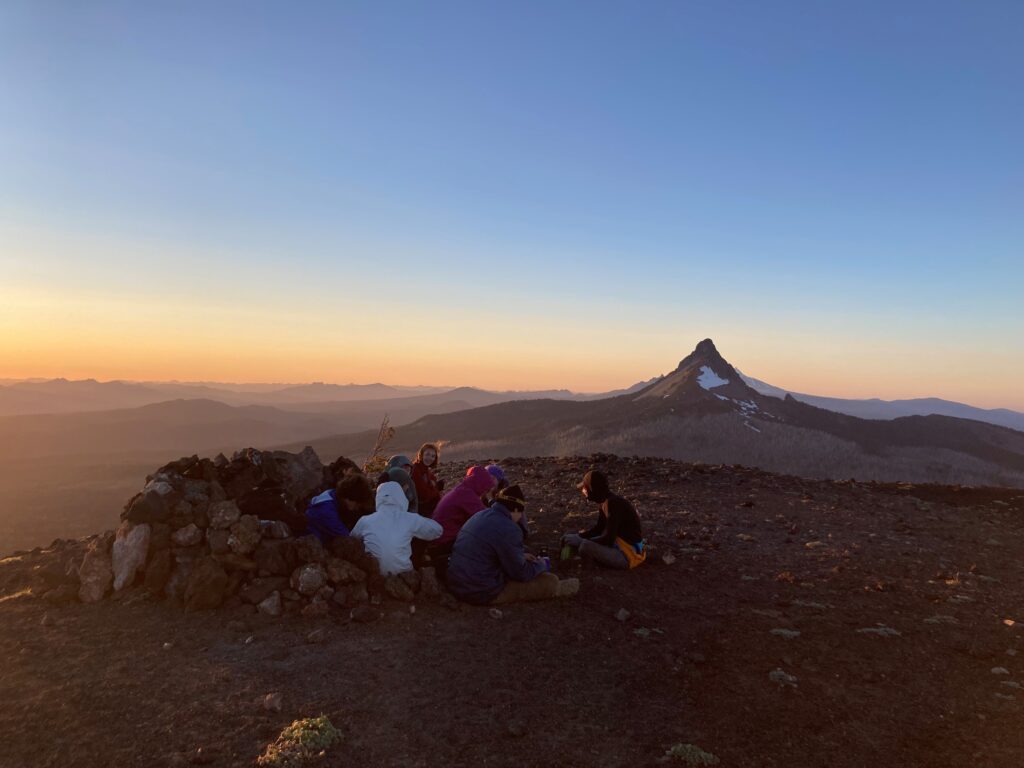 The group of hikers rest next to a stone pile as the sun sets over the horizon.