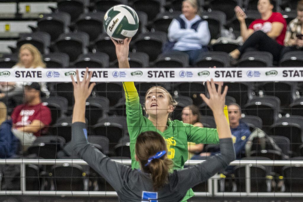 Lynden's Joslyn Sipma (5) soars to tip the ball over the net against a Pullman blocker with their arms raised and ready on the other side of the net.