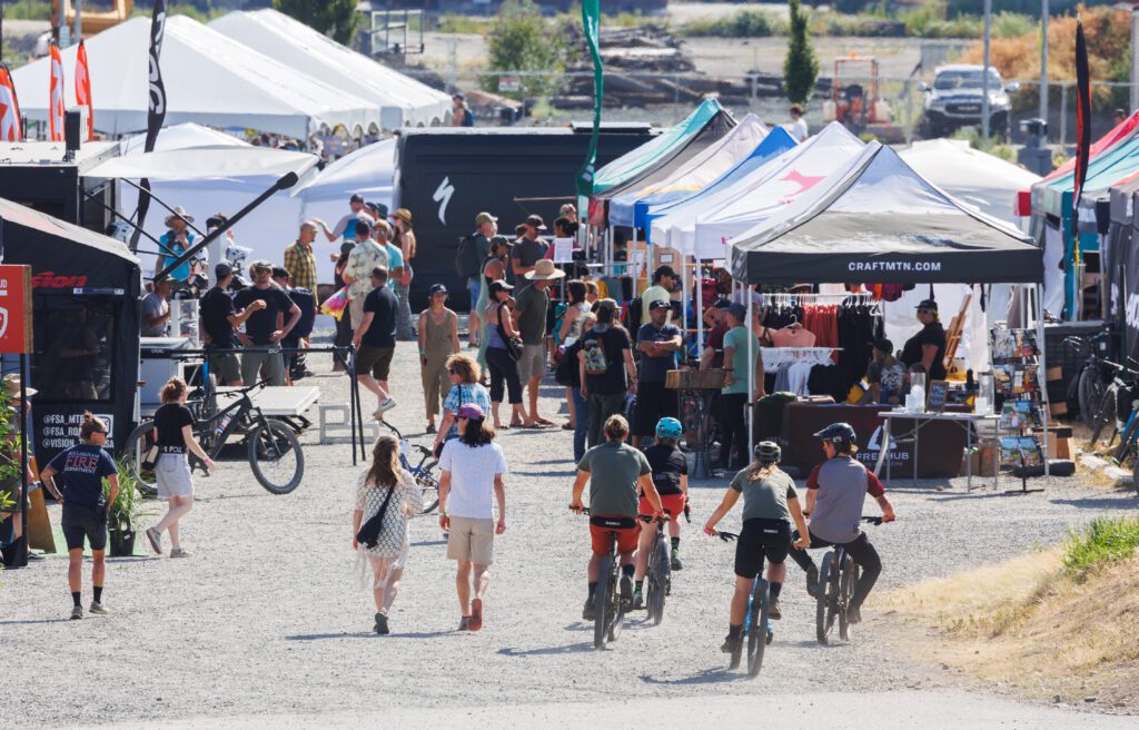 Riders slow down near the vendor area where groups of attendees look around and chat.