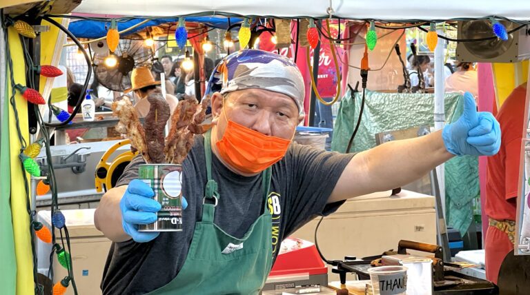 Night Market vendor holding up meat skewers in a can as he gives a thumbs up with his other hand.