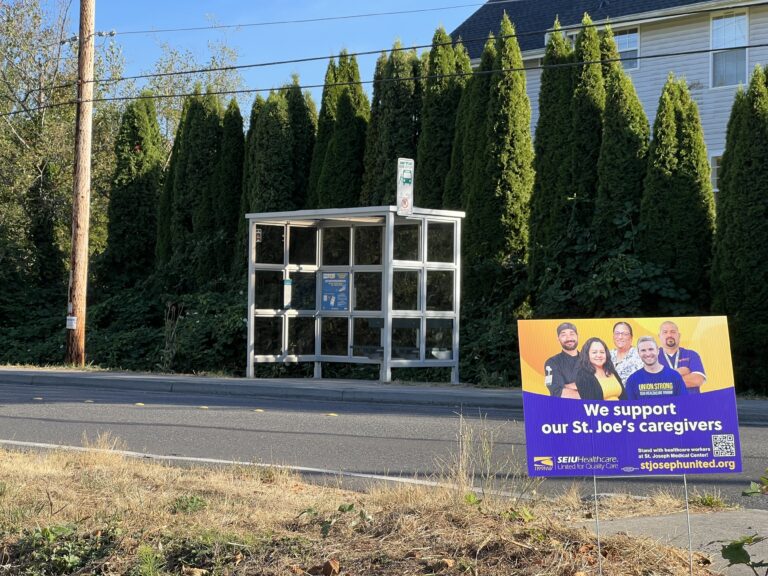 A bright sign with nurses and other healthcare workers supporting PeaceHealth St. Joseph Medical Center stands near a bus stop on Donovan Avenue.