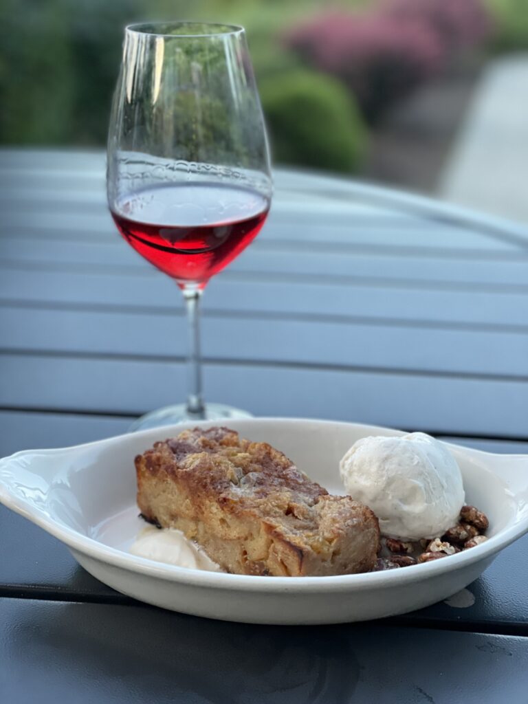 Bourbon bread pudding is served warm with candied pecans and vanilla ice cream next to glass of red wine.