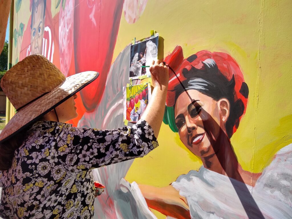 Mattix wears a sunhat as she paints with references taped to the side of the mural.